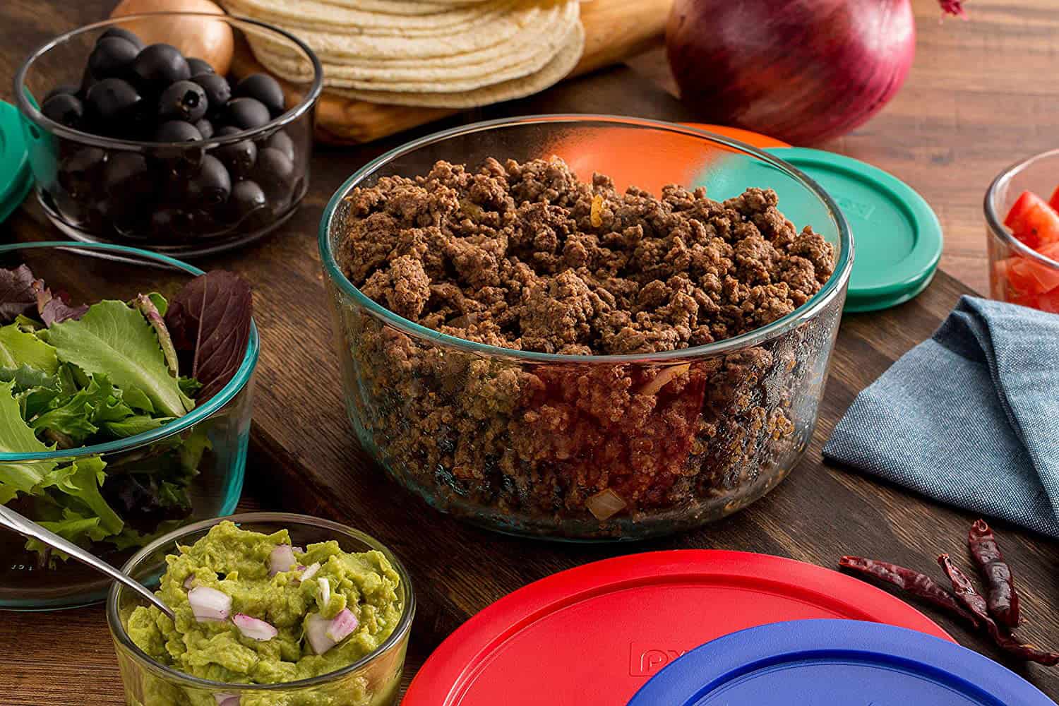 A collection of glass food storage containers with ground meat, lettuce, olives, and guacamole next to plastic lids and tortilla shells.