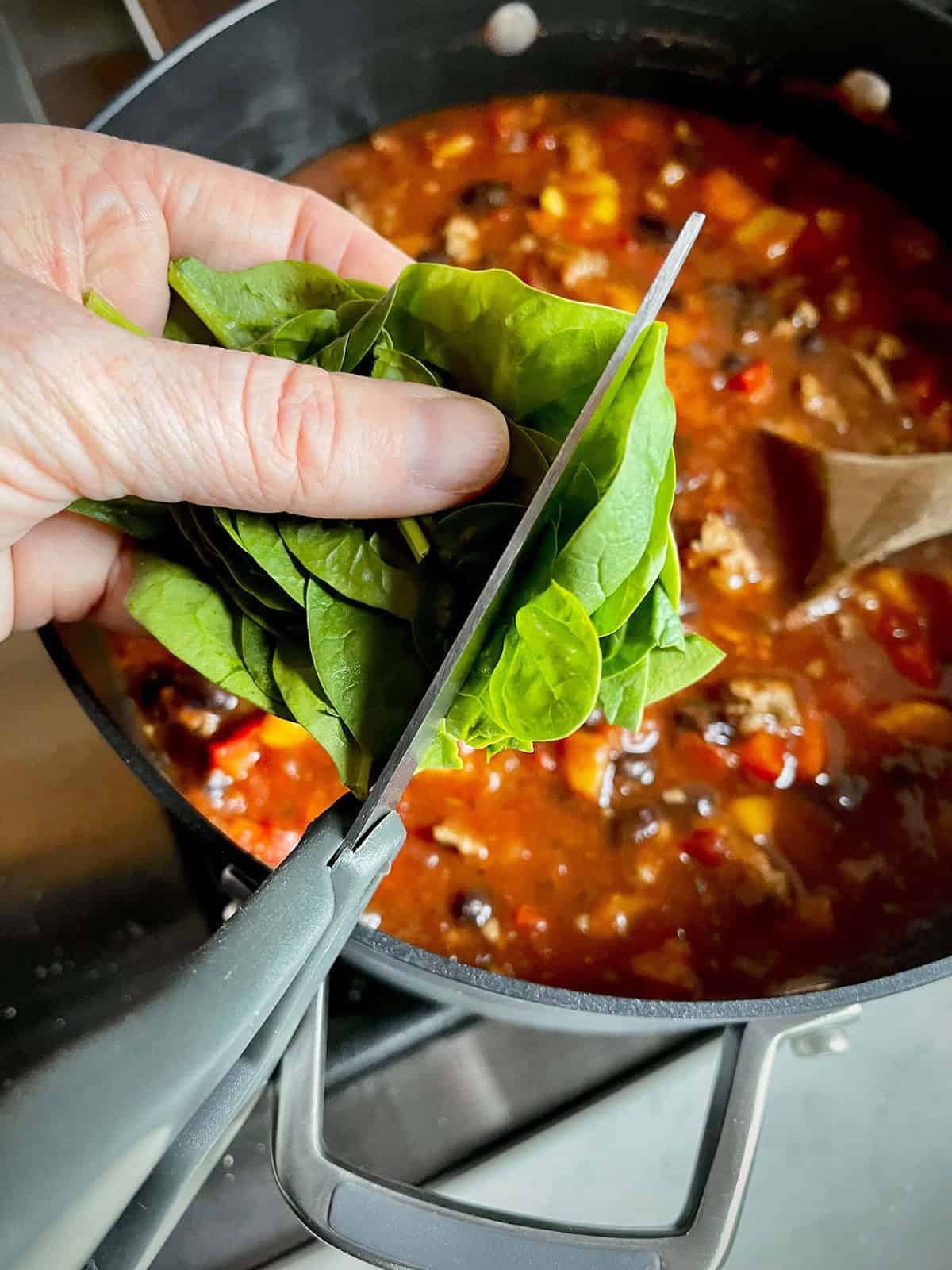 A stovetop pot of turkey taco soup with fresh spinach being added. The baby spinach is being diced with scissors.