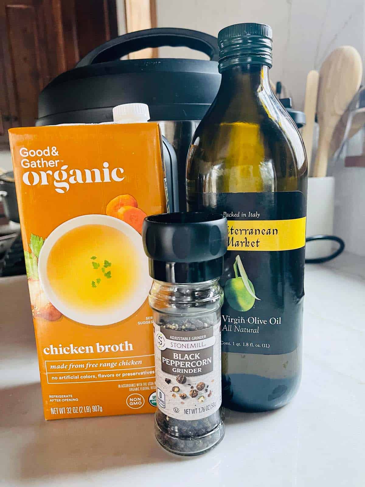 Ingredients for cooking chicken to freeze sitting in front of an Instant Pot; including chicken broth, olive oil, and pepper.